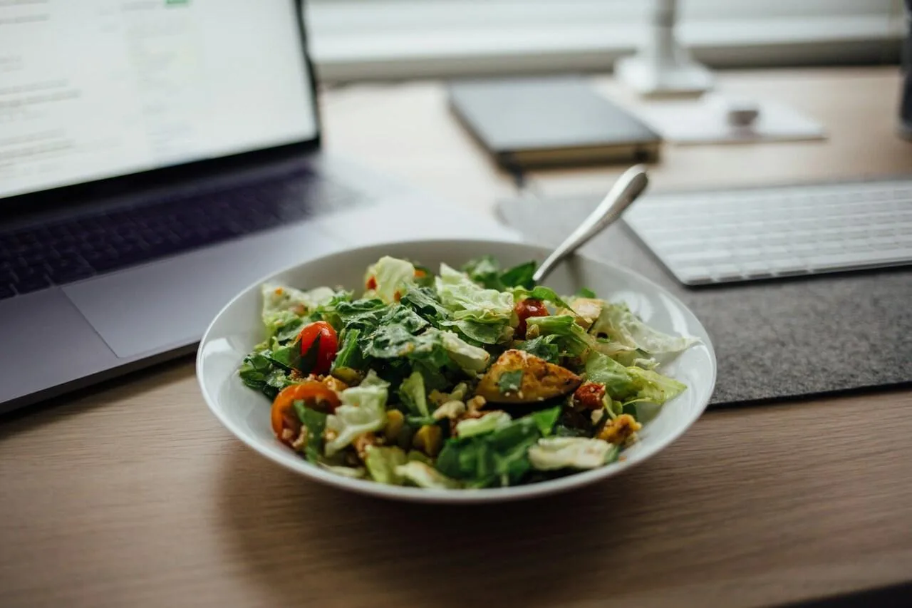 piatto di insalata sana sopra a una scrivania di lavoro davanti a un laptop, una tastiera e un quaderno indicando come mangiare sano quando si ha poco tempo