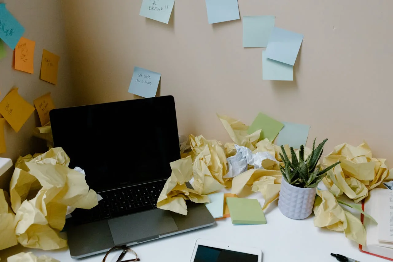 scrivania di lavoro al computer sommersa di appunti post-it e fogli di carta da buttare indicando stanchezza cronica nel lavoro